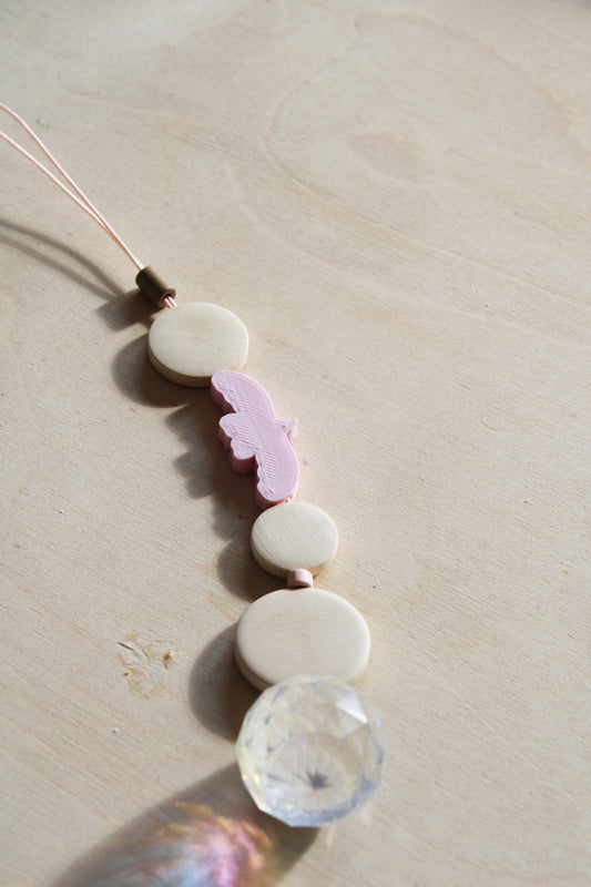 Suncatcher mit einer großen Kristallkugel, Holzperlen, Keramikperlen und 3D-Druck Perle in Form eines Vogels in einem zarten rosa fotografiert auf hellem Holz.