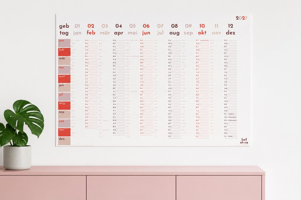 Wandkalender an einer weißen Wand. Darunter steht eine rosa Kommode. Auf dieser steht eine Pflanze in einem hellgrauen Topf.